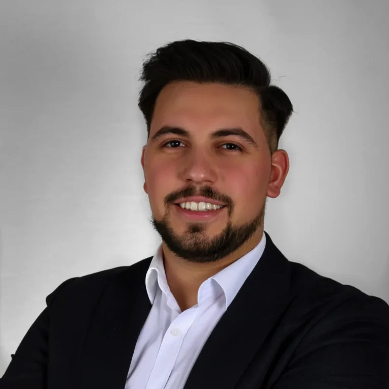 Profilfoto Erkut Akarsu Junger Mann mit Bart und kurzen Haaren in einem Anzug mit verschränkten Armen.